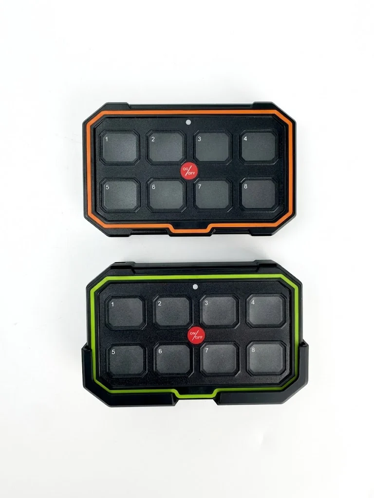 Two RuggedRay 8-gang wireless remote switch panels, one with an orange surround and one with a green surround, each with eight numbered buttons and a central on/off button